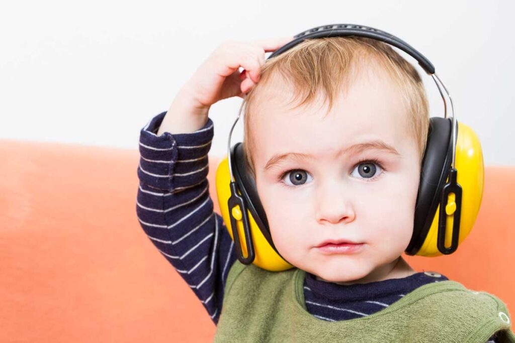 A toddler scratches their head while wearing yellow noise-blocking headphones.