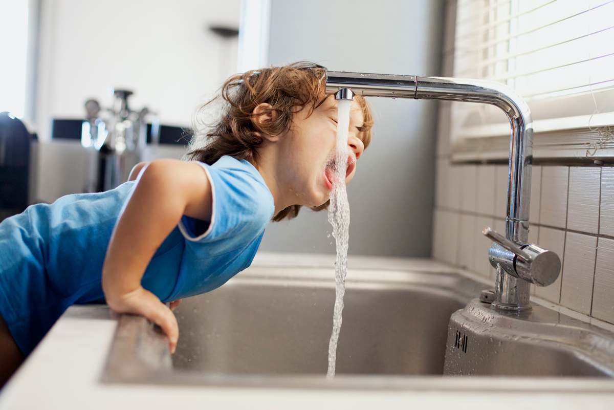 A child leans over a sink to drink water out of a kitchen tap.