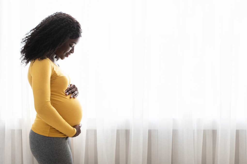 A heavily pregnant person in a yellow shirt paces in front of a window.