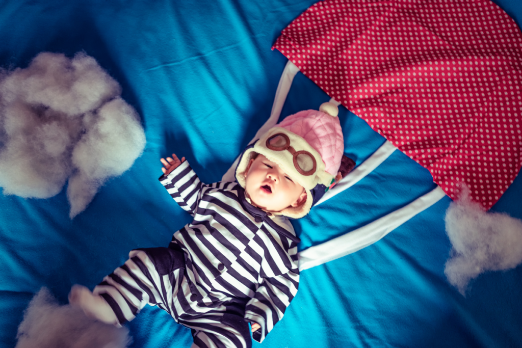 A baby on a blue sheet wears a pretend parachute and skydiving goggles.