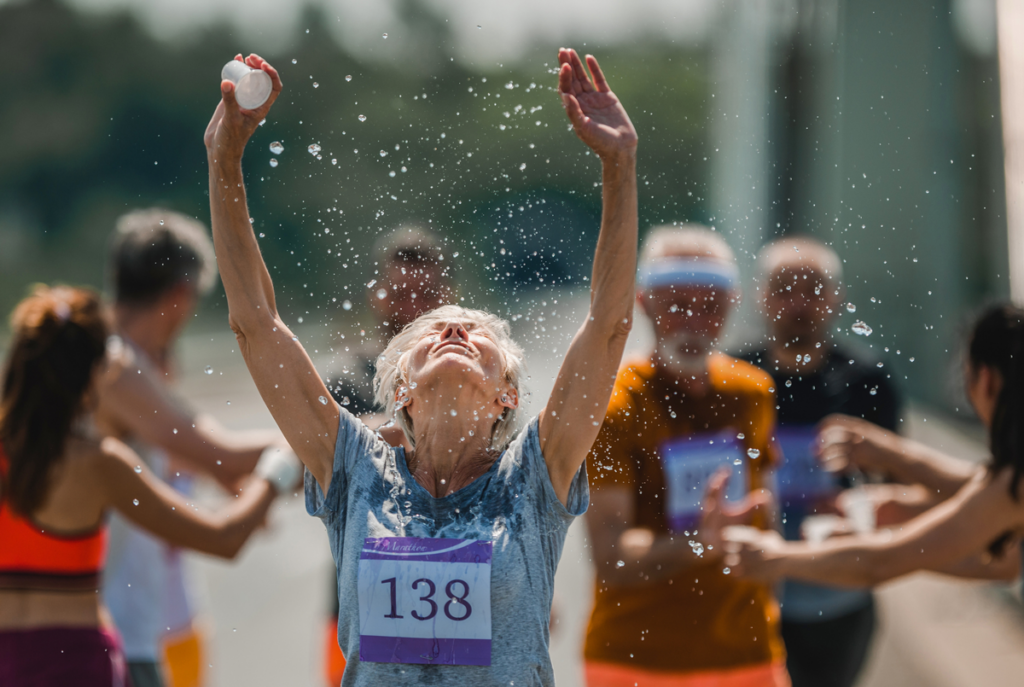 An older running in a marathon tosses water on herself as she crossed the finish line, arms raised.