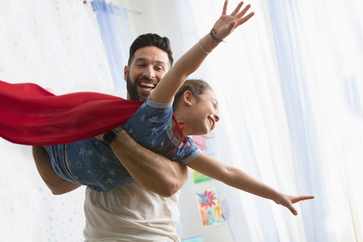 A parent holds up a child, dressed as a superhero, as the child flies through the air.