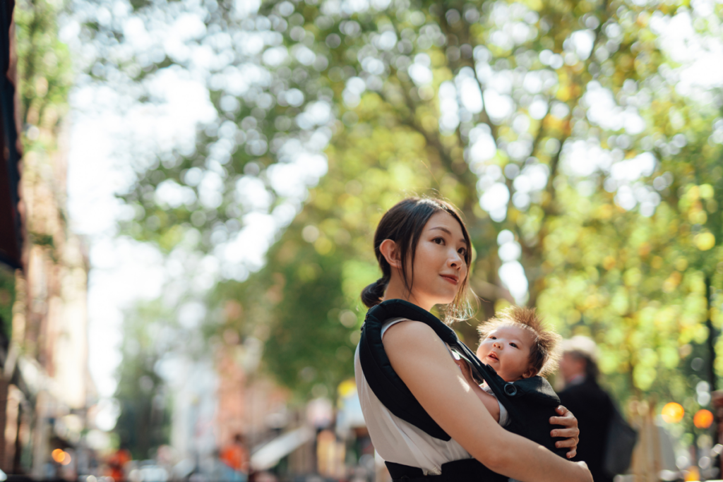 A new parent smiles while holding their baby in an infant carrier outdoors.