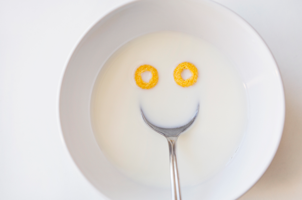 A white bowl of milk contains two pieces of "o"-shaped cereal and a spoon, arranged to look like a smiley face.