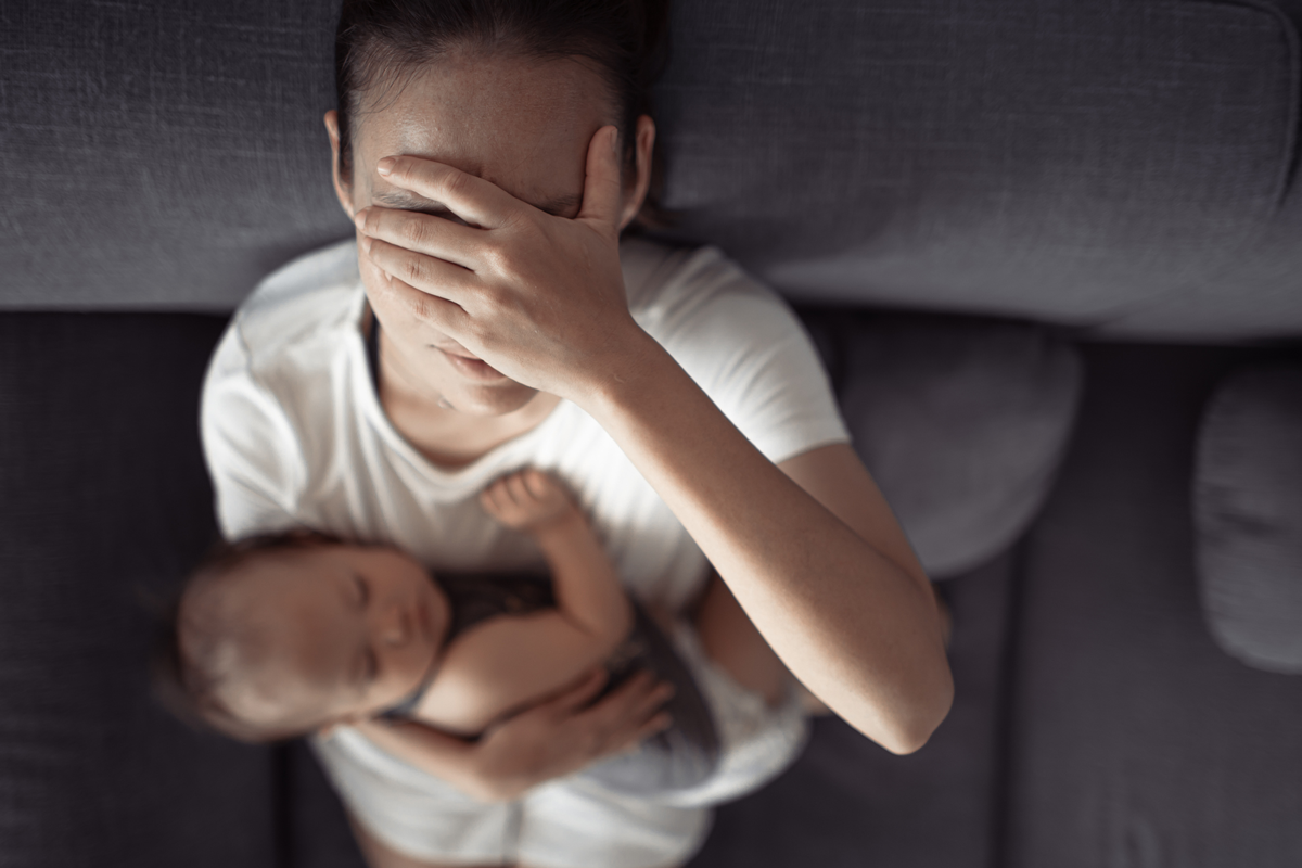 A new parent covers their face in sadness as they sit on a couch holding a baby.