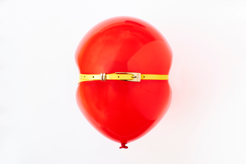 A yellow belt is stretched around a full, red balloon that looks ready to pop.