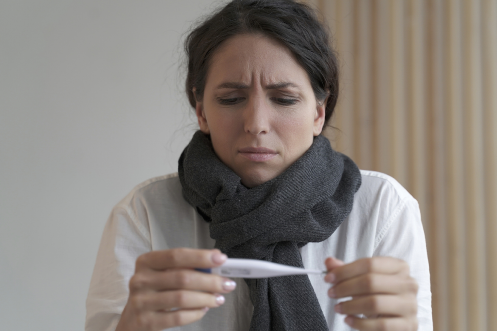A woman wearing a scarf around her neck looks worried as she checks a thermometer.