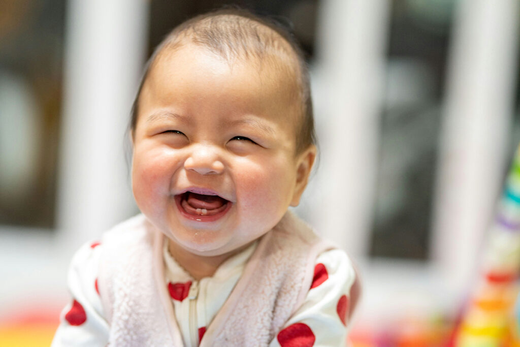 Baby in a sleep sack and printed onesie, smiling big with two bottom teeth.