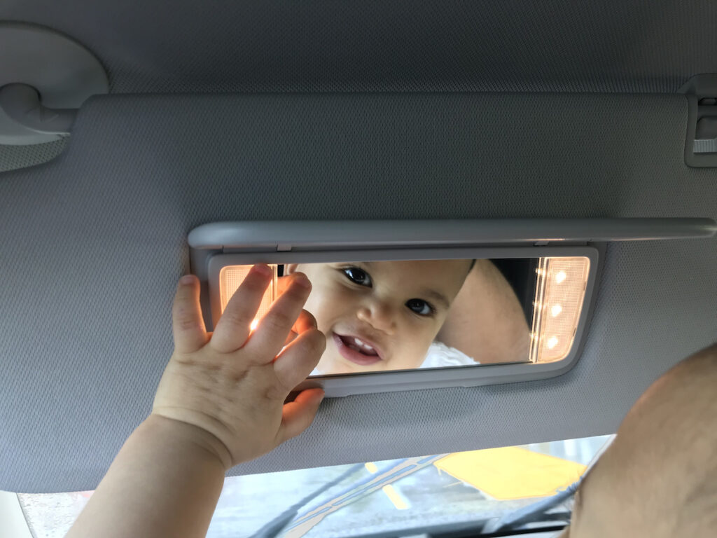 A baby smiles into the review mirror of a car.