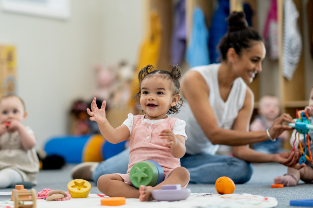 An infant at a daycare smiles while playing with other babies and toys.