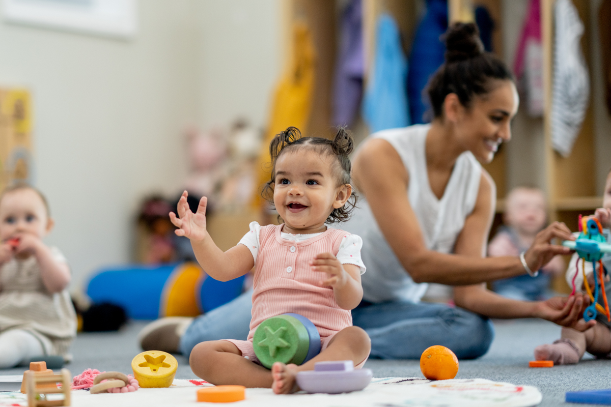 An infant at a daycare smiles while playing with other babies and toys.
