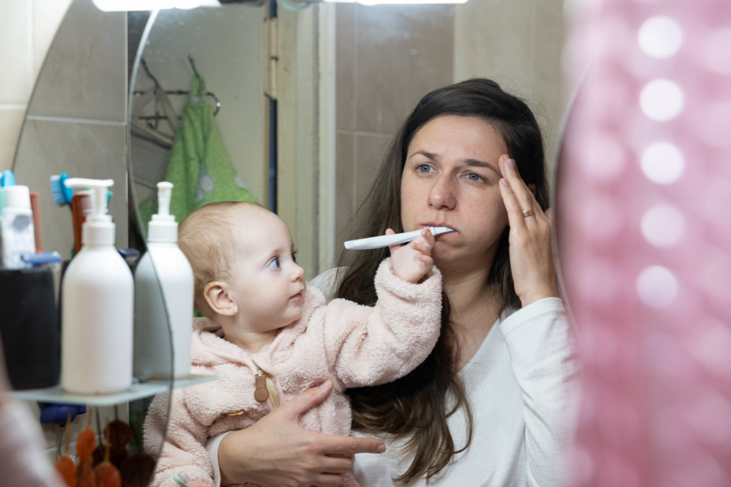 A woman looks into the mirror and rubs her temple as her baby grabs her toothbrush.