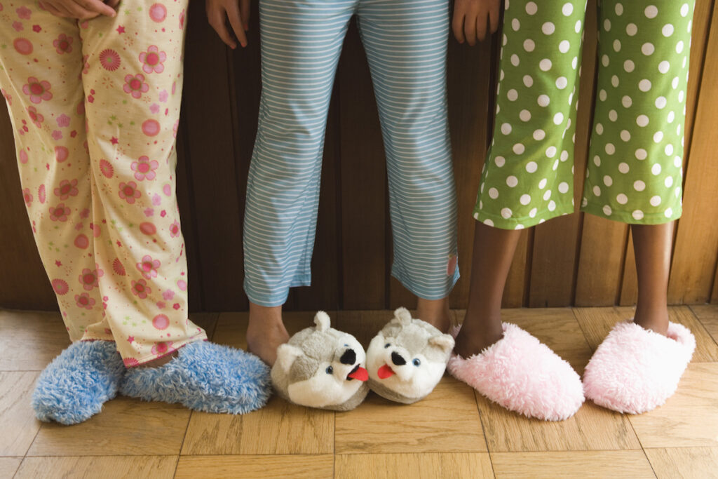 Three pre-teen friends stand next to each other in colorful pajamas and fuzzy slippers at a slumber party.