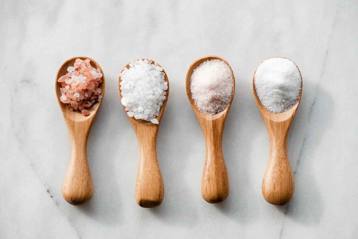 Four tablespoons of various kinds of salt are pictured on a marble background.