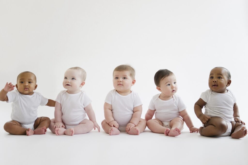 Five babies sit on the floor in a row.