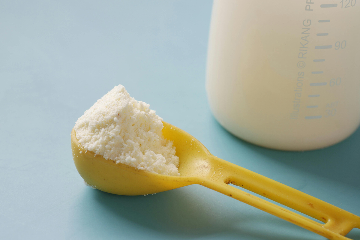 Powdered formula in a measuring spoon, next to a baby bottle.