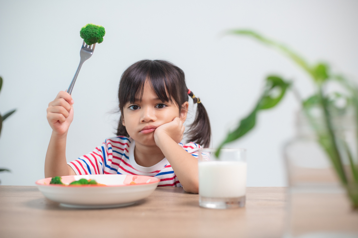 A picky-eater child holds up a stem of broccoli and frowns.