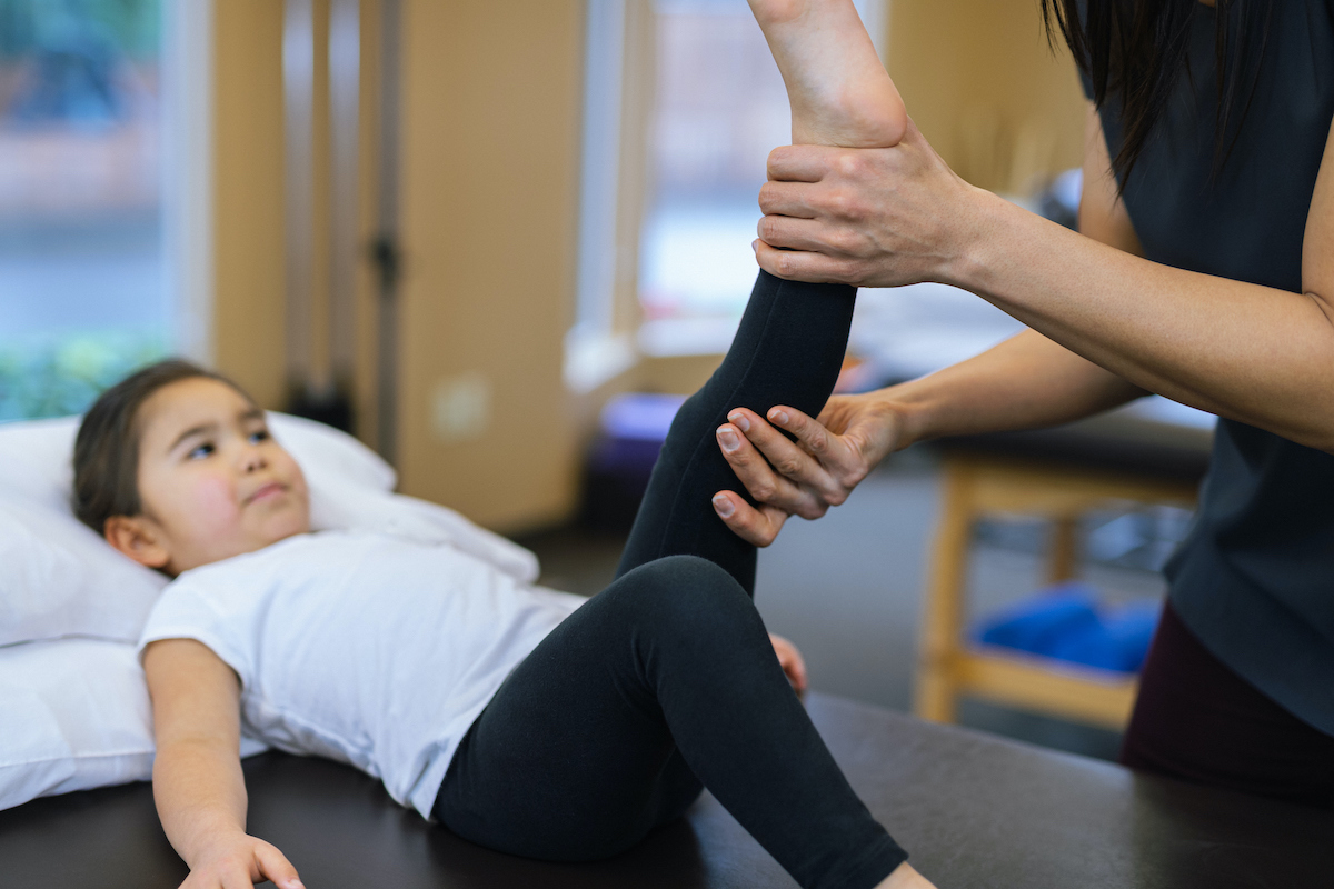 Child (a young girl) lays on a massage bed while female chiropractor lifts her leg