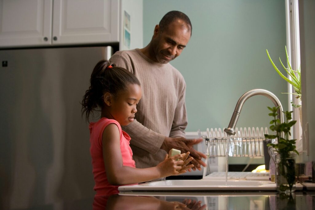 A parent looks on as a child washes their hands at the kitchen sink.