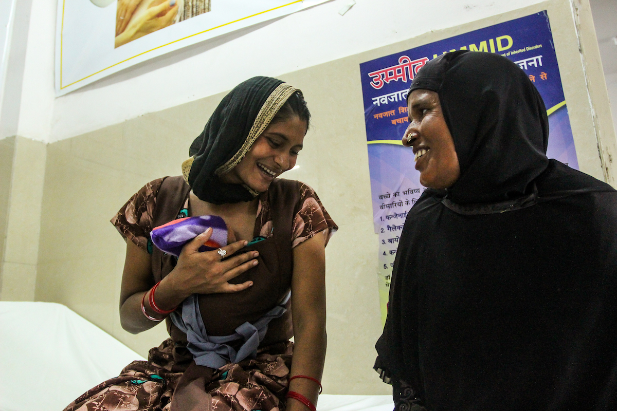 A new parent wearing a baby during skin-to-skin contact smiles and laughs with a visitor.
