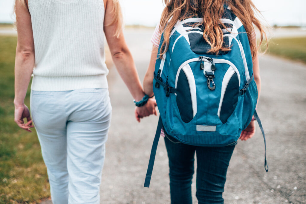 A parent and teen wearing a backpack walk away from the camera.