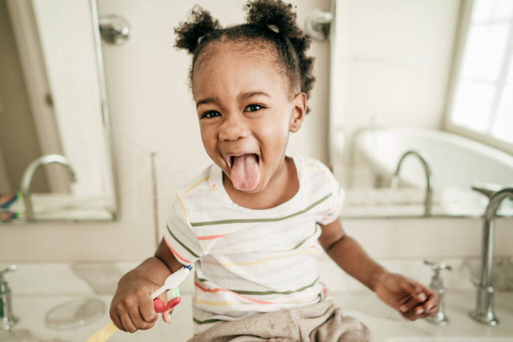 A young child sticks their tongue out while brushing their teeth.