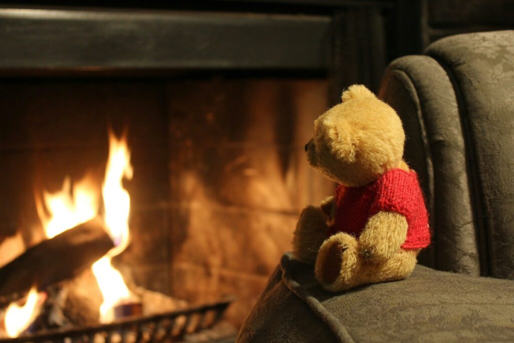 A teddy bear sits on a couch and looks at a fire in a stone fireplace.