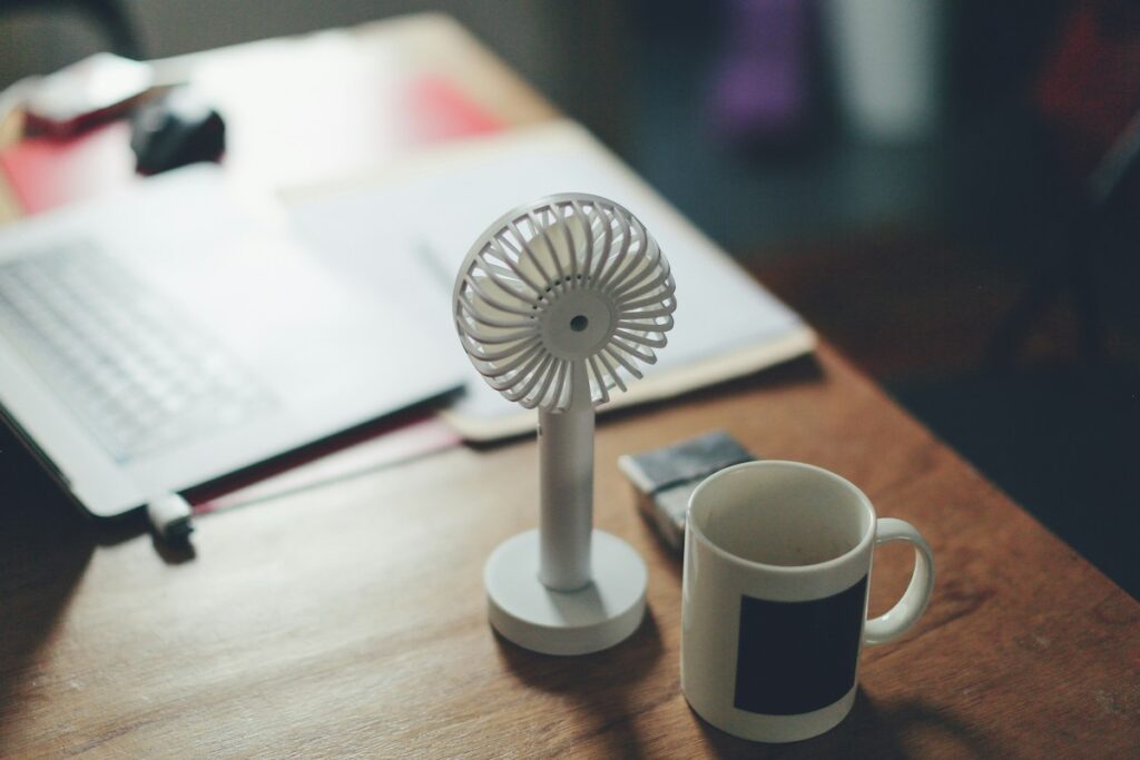 A small fan is pictured next to a cup of coffee and a laptop.