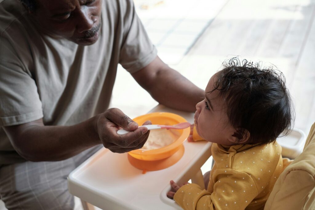 A parent feeds baby food to a baby in a high chair.