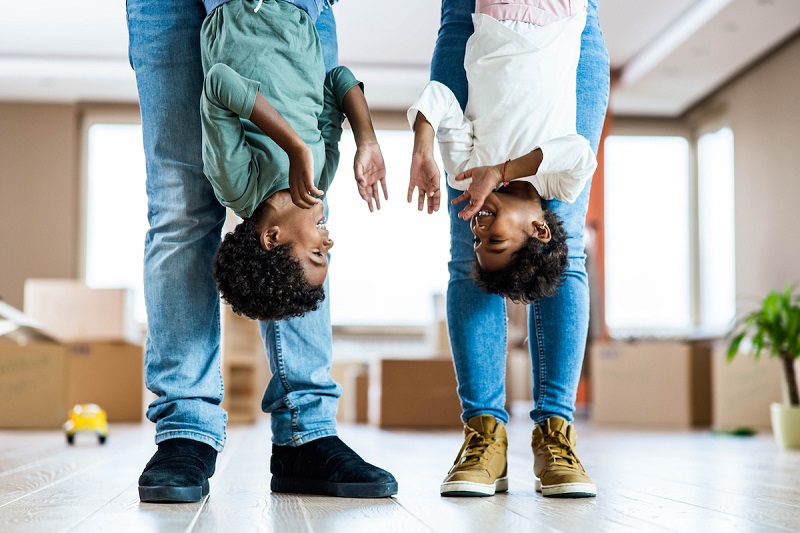 Two kids smile as they are held upside down by their parents, who are shown from the waist down.