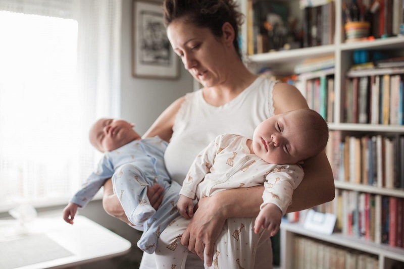 parent holding two babies and standing while looking tired