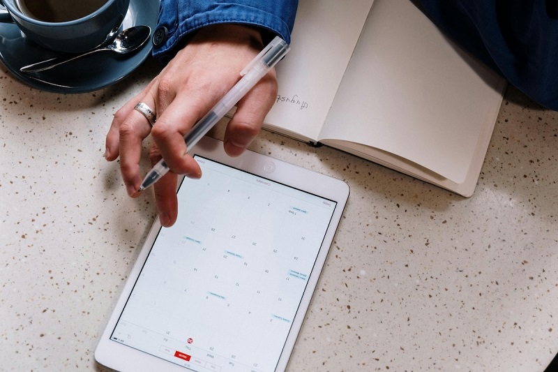 a person writing something on the book and taking information from the tablet