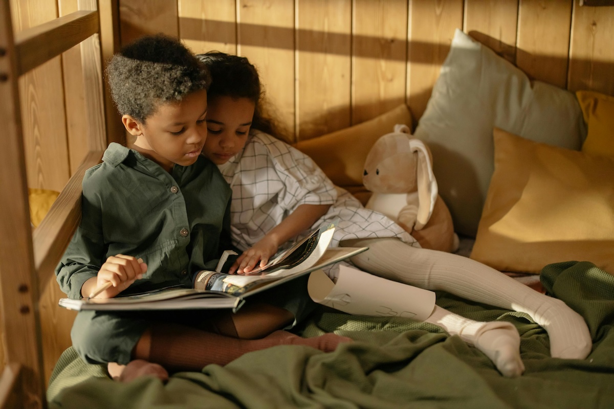 two children siblings sitting on a bed and reading a book together