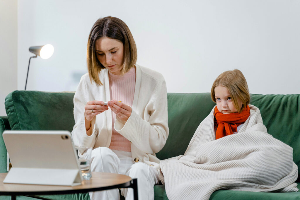 Child wrapped in blanket sitting on couch next to parent who is reading a thermometer