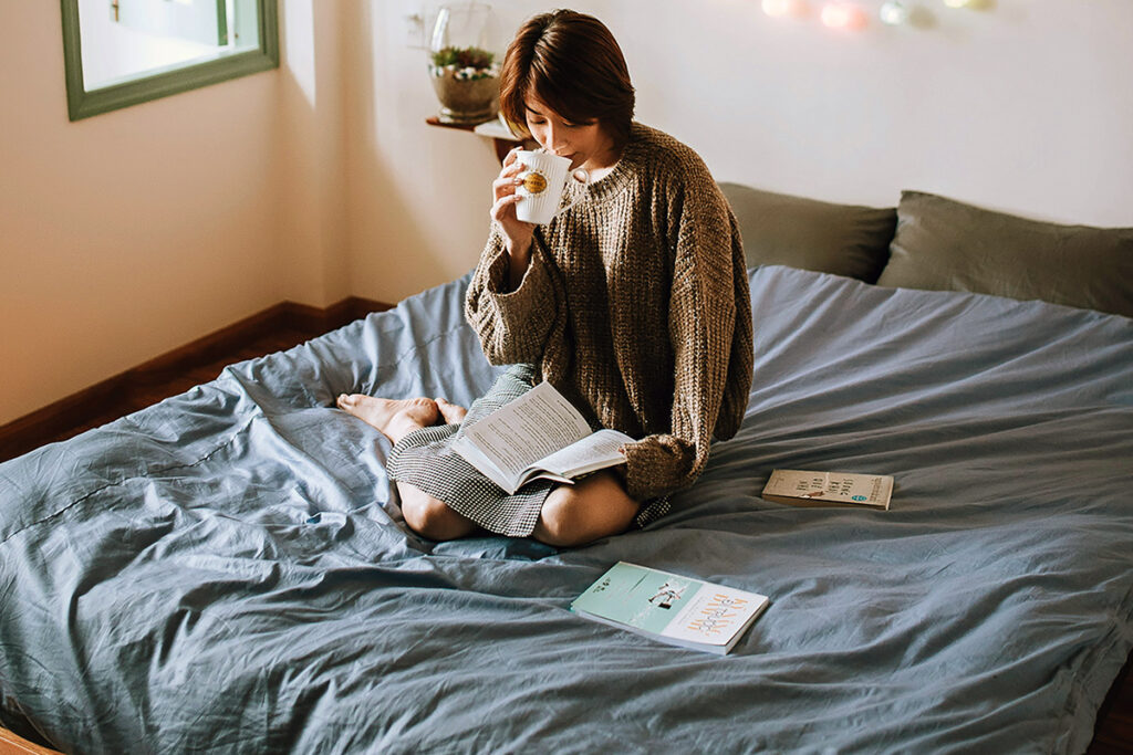 an adult sitting on bed reading book while drinking tea