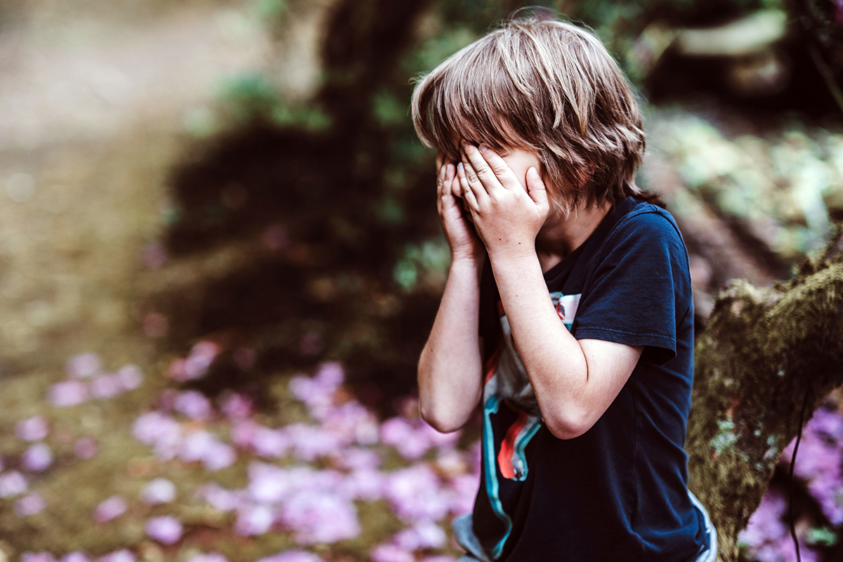 A kid closing his eyes outdoor with trees and flowers in the background