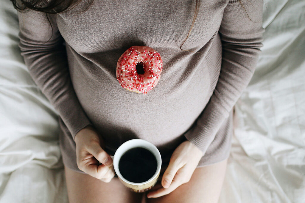 A pregnant person wearing grey dress with donut on the belly and holding coffee