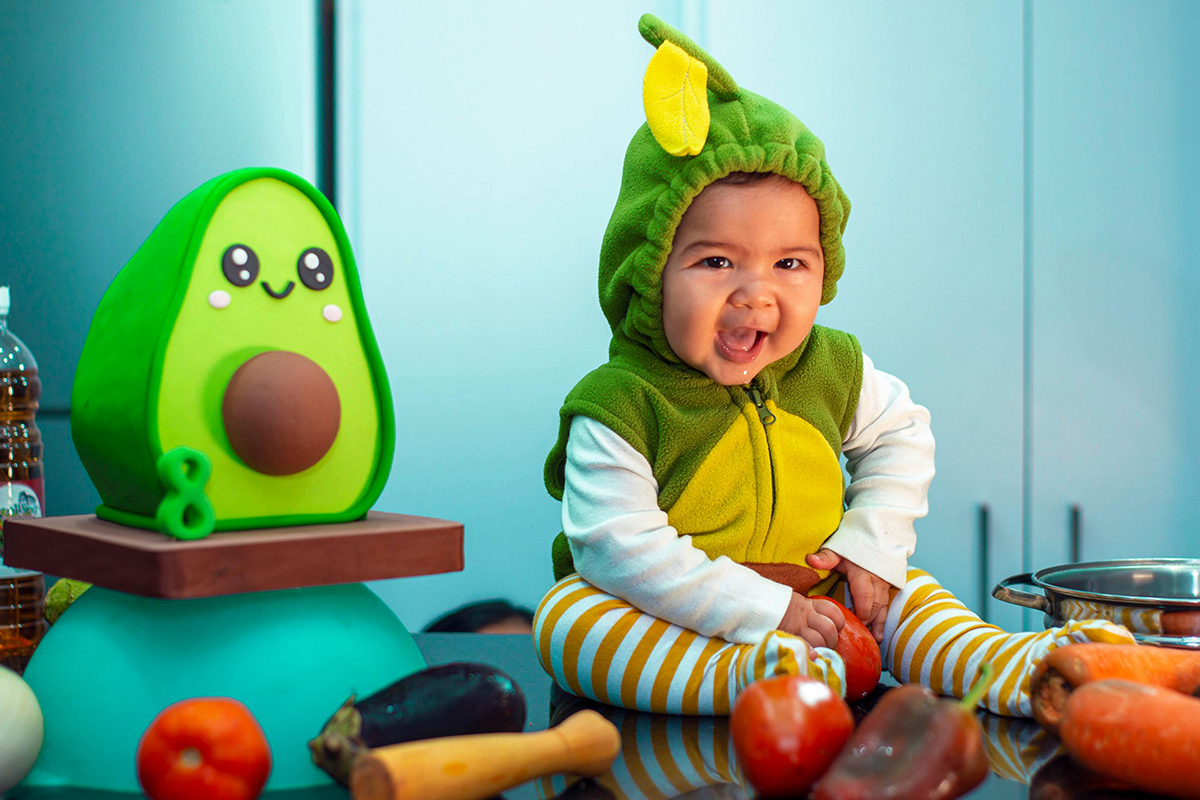 A baby in an avacado costume sitting next to a avacado soft toy