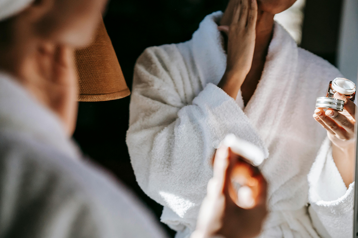 A person in a white bath robe looking into the mirror and touching the face.