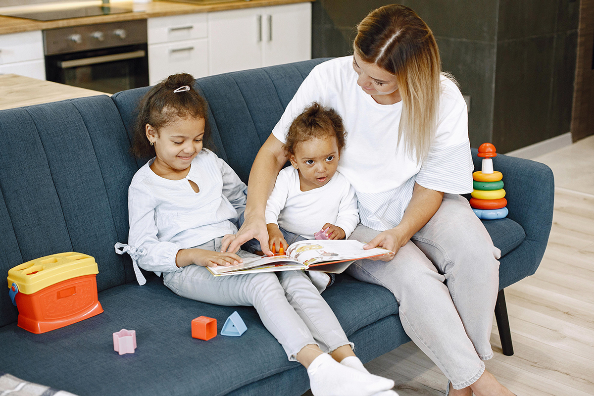 A parent sitting with two children on a couch and teaching them with a book.