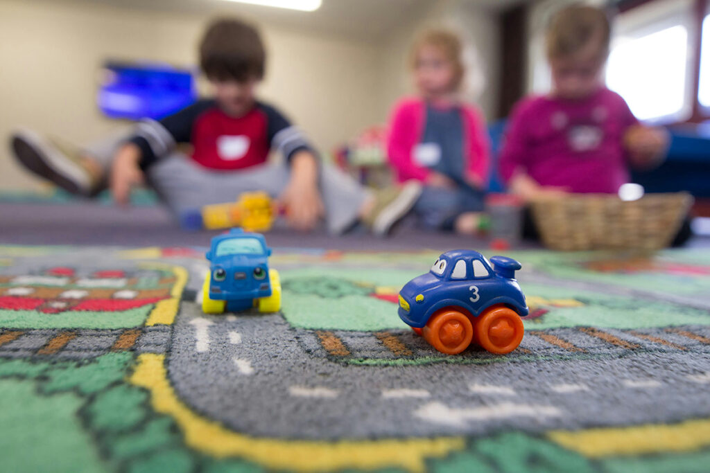 Two toy cars going on a race mat with three kids in the blurred background.