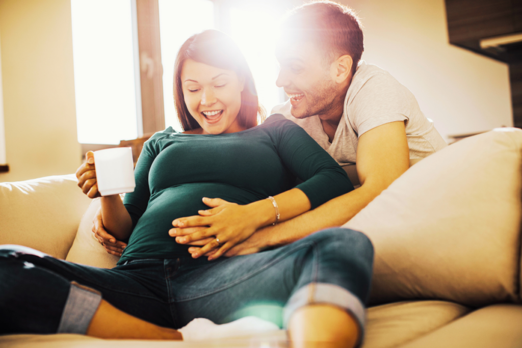 A couple sitting together on the sofa holding a pregnant belly and smiling