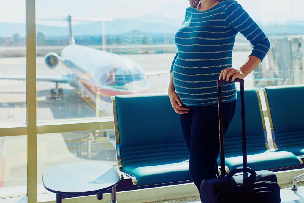 Pregnant person standing with one hand on a suitcase and one hand on belly with an airplane outside the window