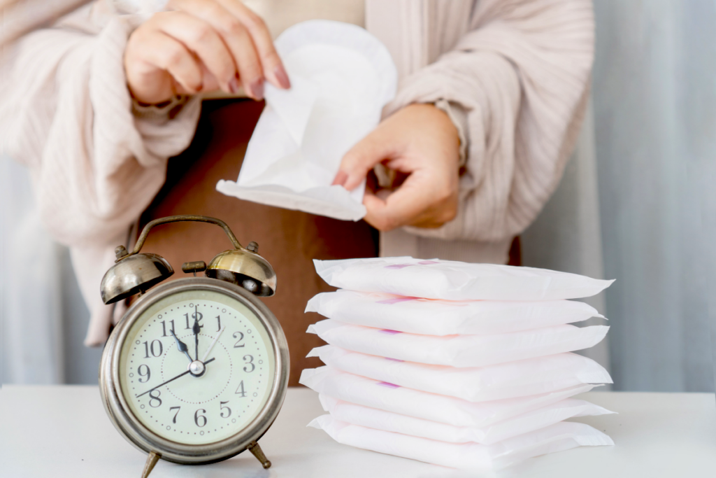 A clock in the foreground with a woman opening a sanitary napkin in the background.