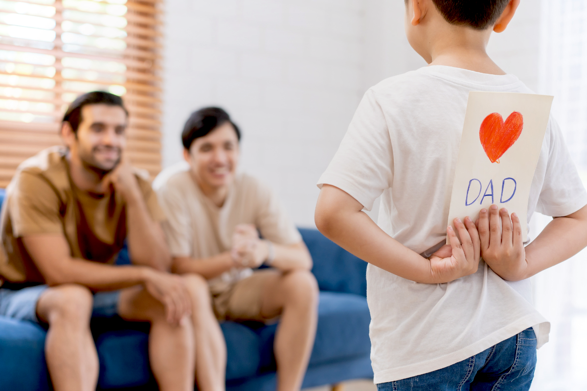 Parents sitting on the couch and looking at their kid holding a card at the back.