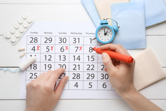 Hands crossing off dates in a calendar with a clock, sanitary napkins, a tampon and a pill strip by the side.