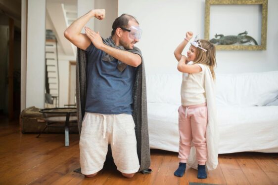 A parent and a child wearing a cape made of blanket, goggles and flexing their arms