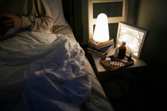 Night stand with a lamp and pills by the side of bed.