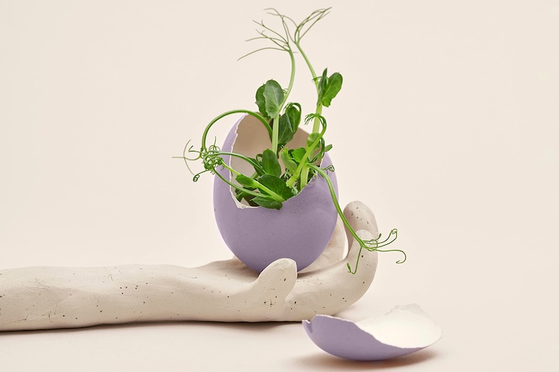 sculpture of a hand holding a lavender egg with the top broken off and pea shoots growing out of it