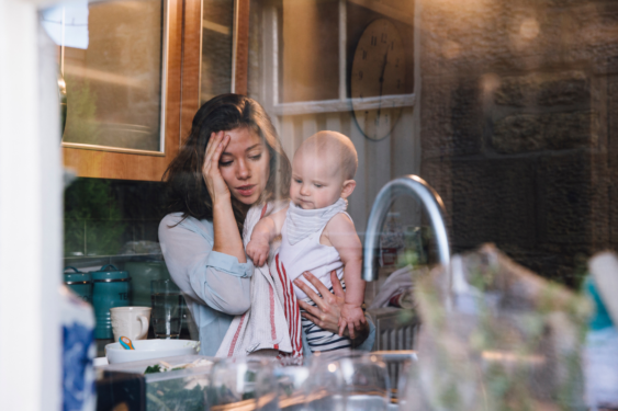 A parent in a kitchen looking stressed with a baby.